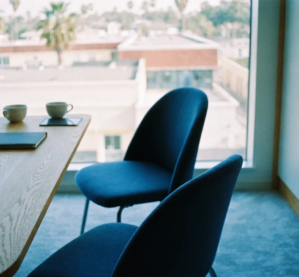 An empty meeting table by a window with two navy chairs, a notebook and two coffee cups